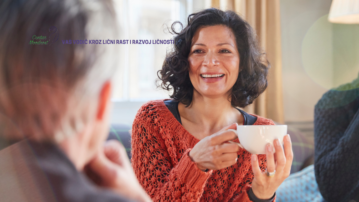 Nasmejana žena crne kose pije kafu iz bele šoljice.